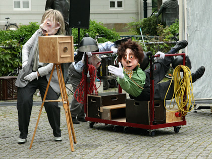 Blaumeier Atelier - Drei Schauspieler stehen auf einem Platz draußen. Sie tragen große bemalte Maskenköpfe aus Pappmaché. Einer steht hinter einer altmodischen Kamera aus Holz. Die beiden anderen bringen im Hintergrund einen Wagen mit Scheinwerfern und Kisten.
