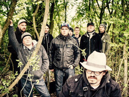Kosmonovski - Neun Musiker stehen in einem überwucherten grünen Wald. Einer hat einen weißen Hut und eine Brille auf. Sie schauen in die Kamera.