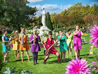 Mademoiselle Orchestra - Acht Musikerinnen in bunten Kleidern stehen auf einer Wiese. Sie spielen auf Saxophonen, Gitarren und Trompeten. Im Hintergrund sieht man eine weiße Statur einer Frau in einer Robe.