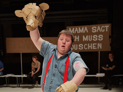 Meine Damen und Herren - Gans der Bär - Ein Schauspieler mit Down-Syndrom steht auf einer dunklen Bühne. Auf einer Hand trägt er eine Bärenmaske aus brauner Pappe, auf der anderen Hand eine braune Papp-Tatze. Im Hintergrund sieht man ein Schild mit weißen Buchstaben, auf dem steht: Mama sagt ich muss duschen.