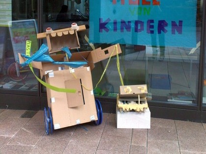 Workshop Puppenbau - Eine Figur aus Pappkartons zusammengebaut steht vor einem Schaufenster. Sie hat Räder und einen weit aufgerissenen Mund mit vier Zähnen. Dahinter hängt ein blaues Plakat auf dem steht: Kunst aus Müll von Kindern