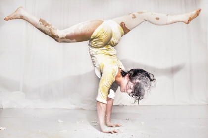 Natalie Reckert - Eine Artistin macht einen Handstand. Die Beine hat sie dabei zur Seite gestreckt. Sie trägt einen gelben Turnanzug und ihr Körper ist mit weißem Puder oder Mehl bedeckt.