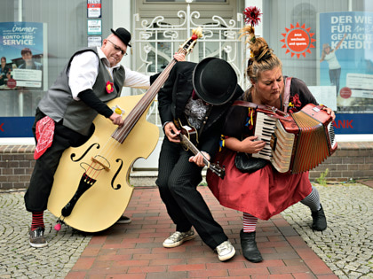 The Bombastics - Die drei Musiker spielen ihre Instrumente auf der Straße vor einem Schaufenster. 