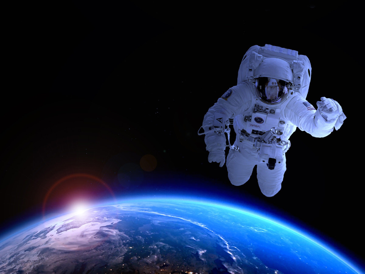 Eine Person in eine weißen Raumabzug schwebt im Orbit. Sie winkt in die Kamera. Unten sieht man ein Stück vom blauen Planeten Erde.