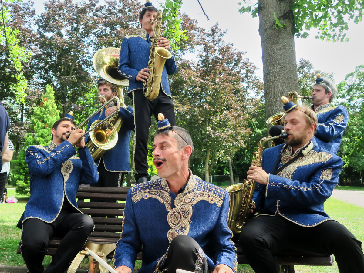 Sechs Menschen mit Blechblasinstrumenten in einem Park. Sie tragen blaue Band-Uniformen mit goldenen Verzierungen.