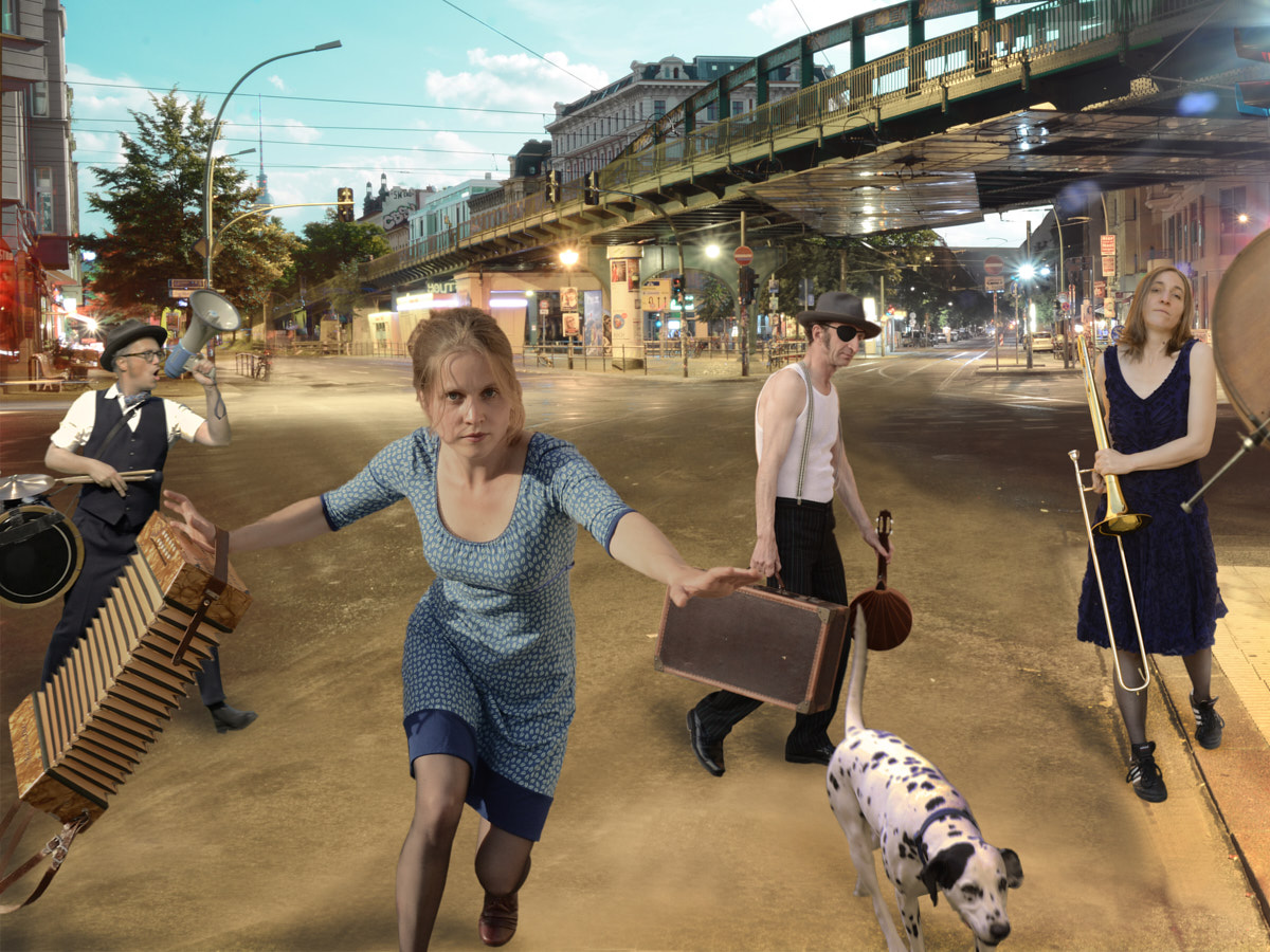 Zwei Musikerinnen und zwei Musiker und ein schwarz-weißer Hund laufen über eine Straßenkreuzung. Sie tragen ihre Instrumente. Hinten sieht man eine Brücke und eine U-Bahn.