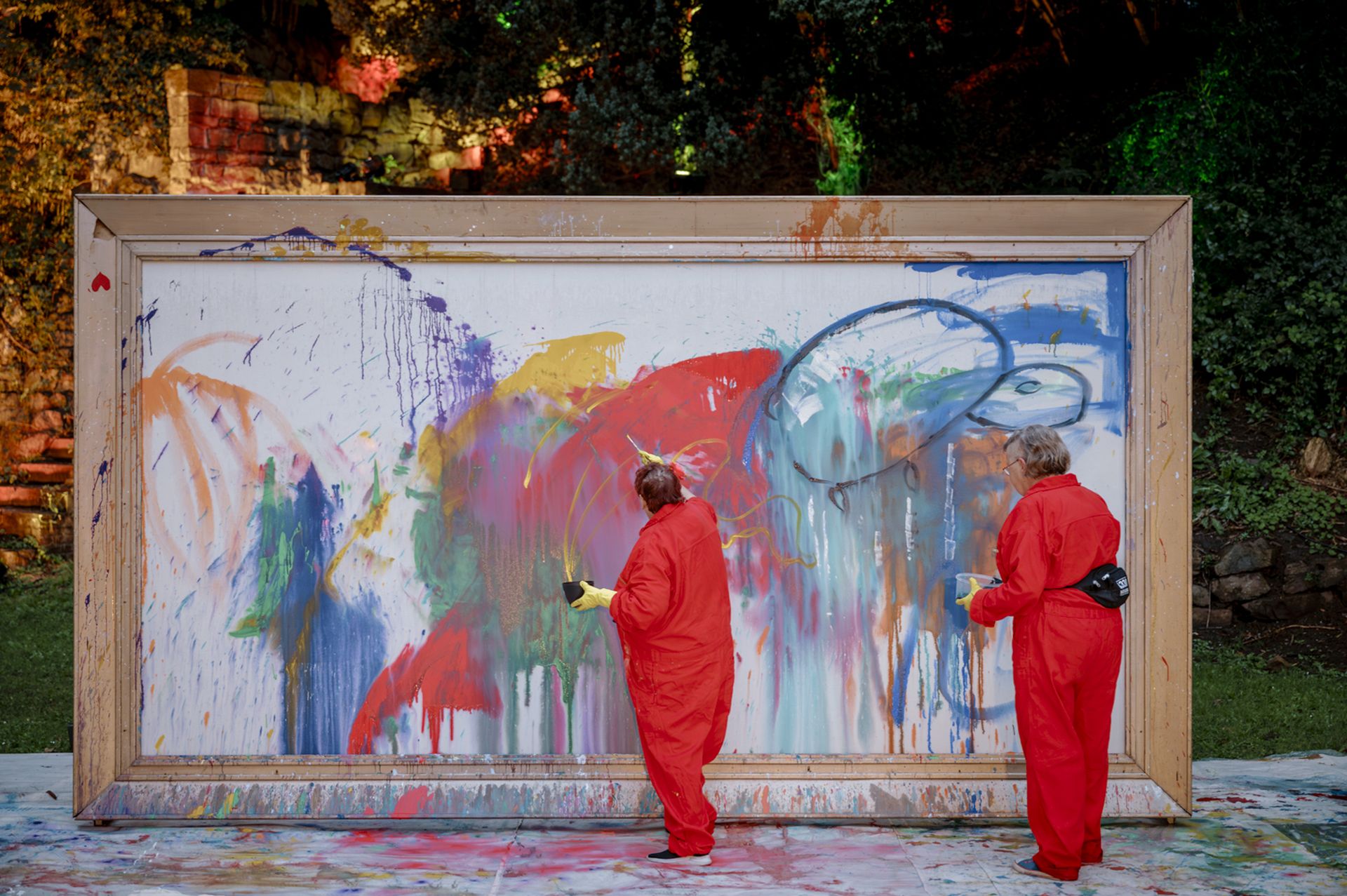 Farbfotografie von zwei Personen vor einer übergroßen Leinwand, die im Freien steht. Die Malenden tragen rote Overalls. Auf der weißen Leinwand sind viele Farbkleckse, in Orange, Rot, Hellblau, Grün, Dunkelblau, Gelb.