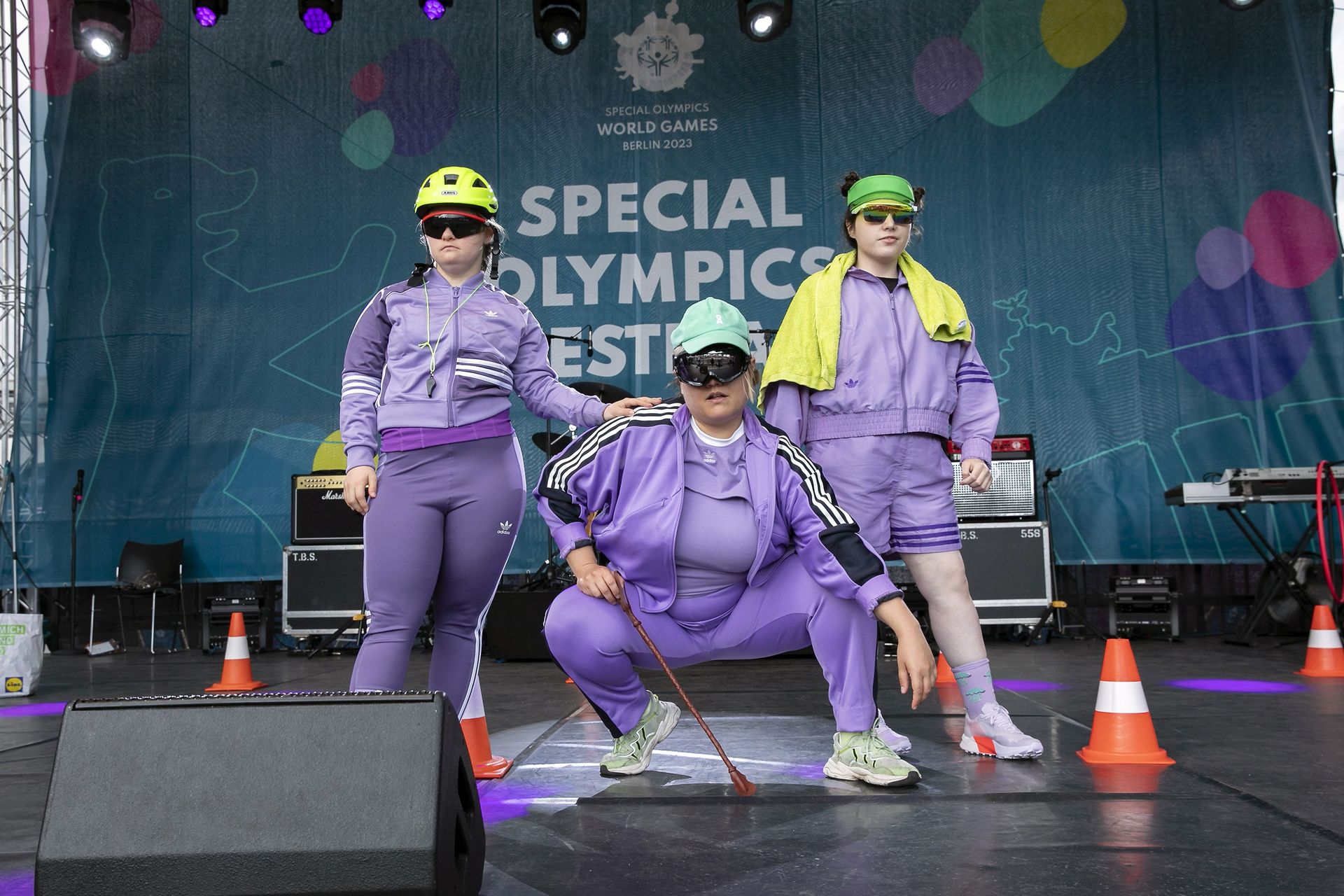 Farbfoto. Drei Personen in lila Trainingsanzügen, Sonnenbrillen und sportlichen Kopfbedeckungen. Die mittlere Person hockt zwischen den anderen. Im Hintergrund hängt der türkise Banner "Special Olympics Festival".