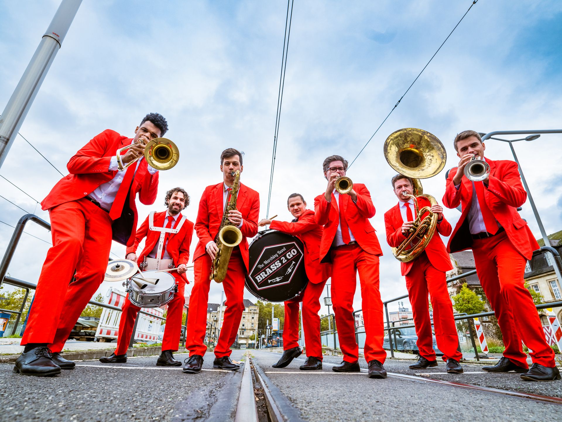 Farbfoto von sieben Musikern in knallroten Anzügen. Sie stehen mit ihren Instrumenten auf Straßenbahnschienen unter freiem Himmel. Dabei sind zwei Trompeten, eine Tuba, eine Posaune, eine Snare, eine Trommel und ein Saxophon.