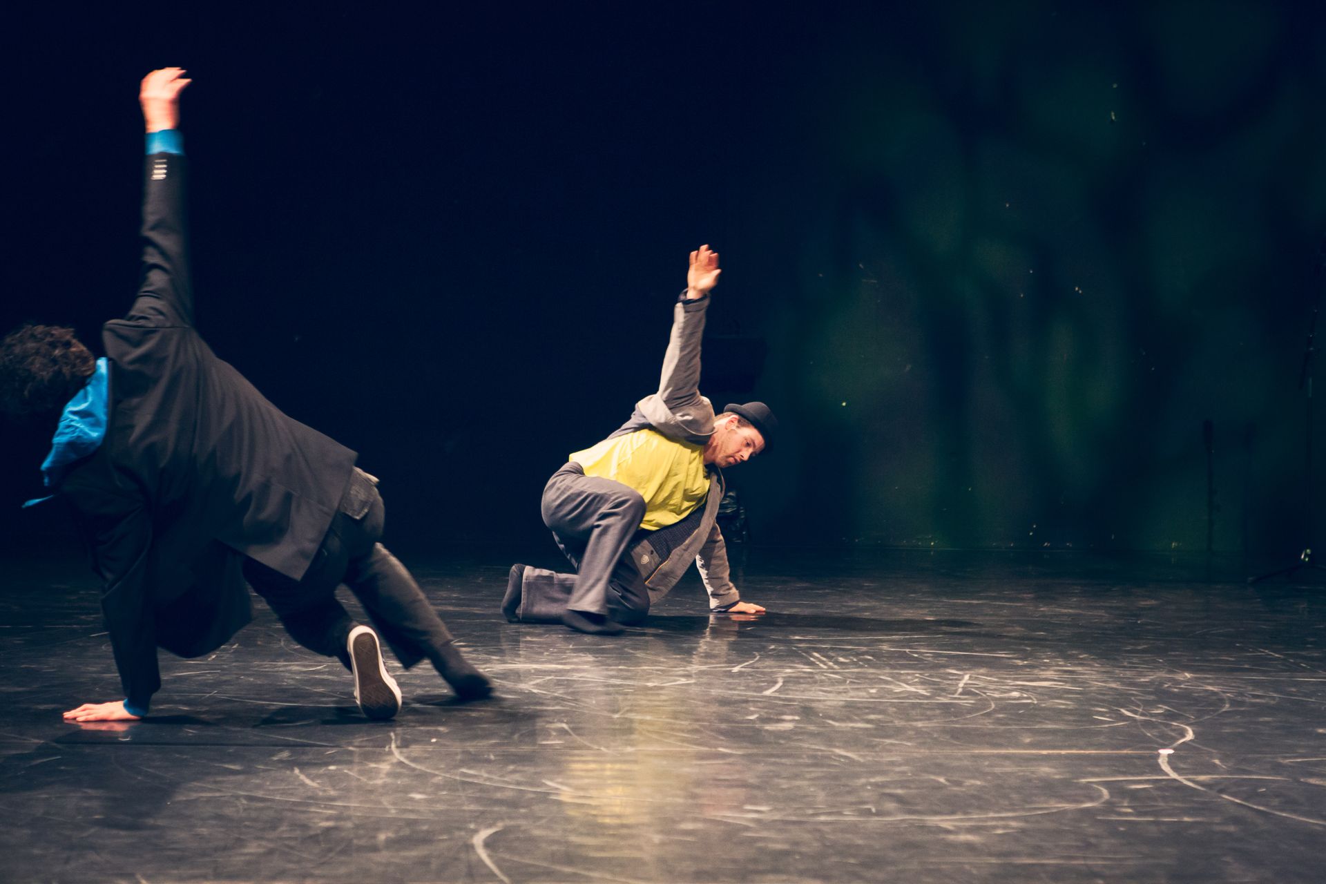 Foto von zwei Performern in der linken Hälfte des Bildes. Sie stützen sich einhändig kniend auf einen Tanzboden, während sie den zweiten Arm gerade über den Kopf strecken. Das Bild vermittelt eine Atmosphäre gemischt aus Breakdance und Parkour.