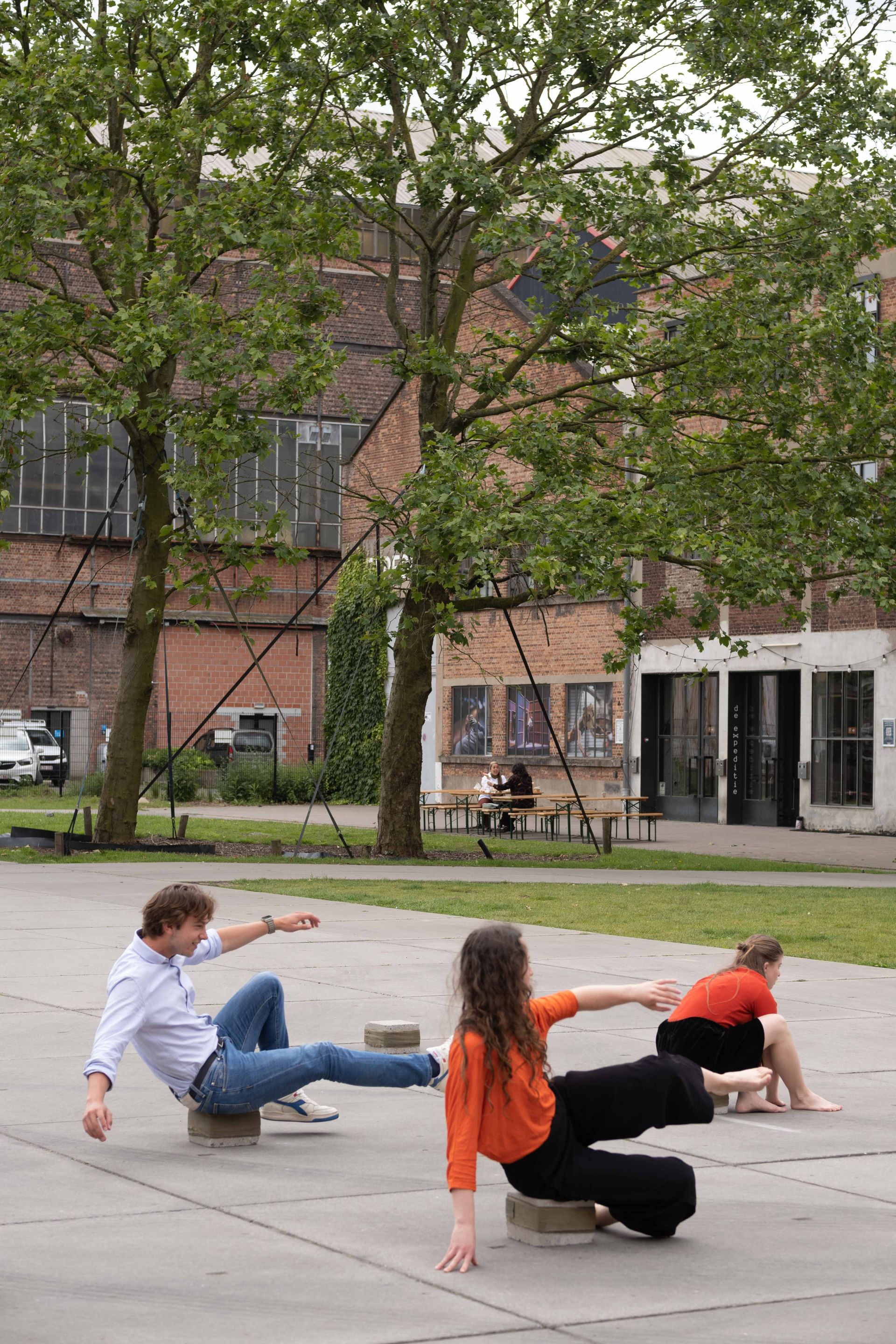 Farbfoto von drei Performer*innen, die auf "Sitzkissen" aus Stein am Boden balancieren, Beine und Arme von sich gestreckt, um das Gleichgewicht zu halten. Im Hintergrund befinden sich Bäume, Bierbänke und zwei Gebäude aus Backstein.