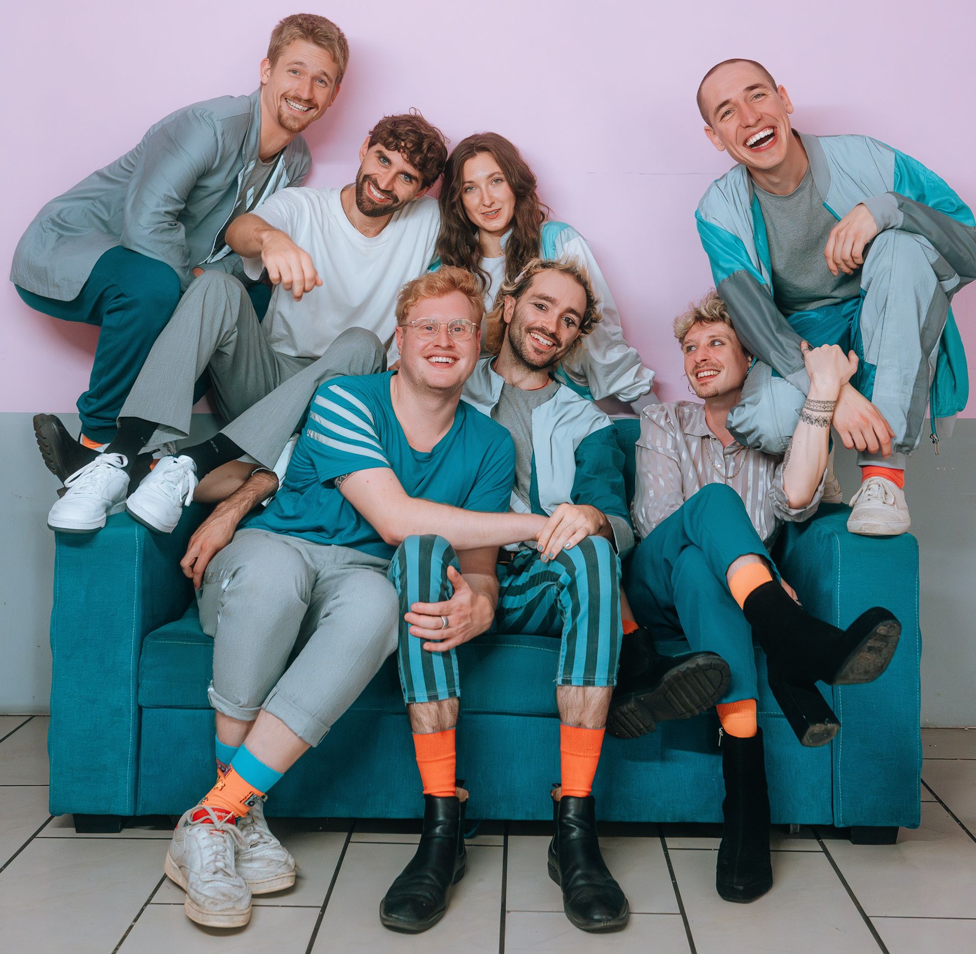 Farbfotografie von sieben Personen, die alle auf ein blaues Sofa arrangiert sind. Dabei stehen zwei Menschen auf den Armlehnen, drei Sitzen auf den Kissen und zwei sitzen auf der Rückenlehne. Alle lachen in die Kamera. Ihre Kleidung ist blau und grau, mit