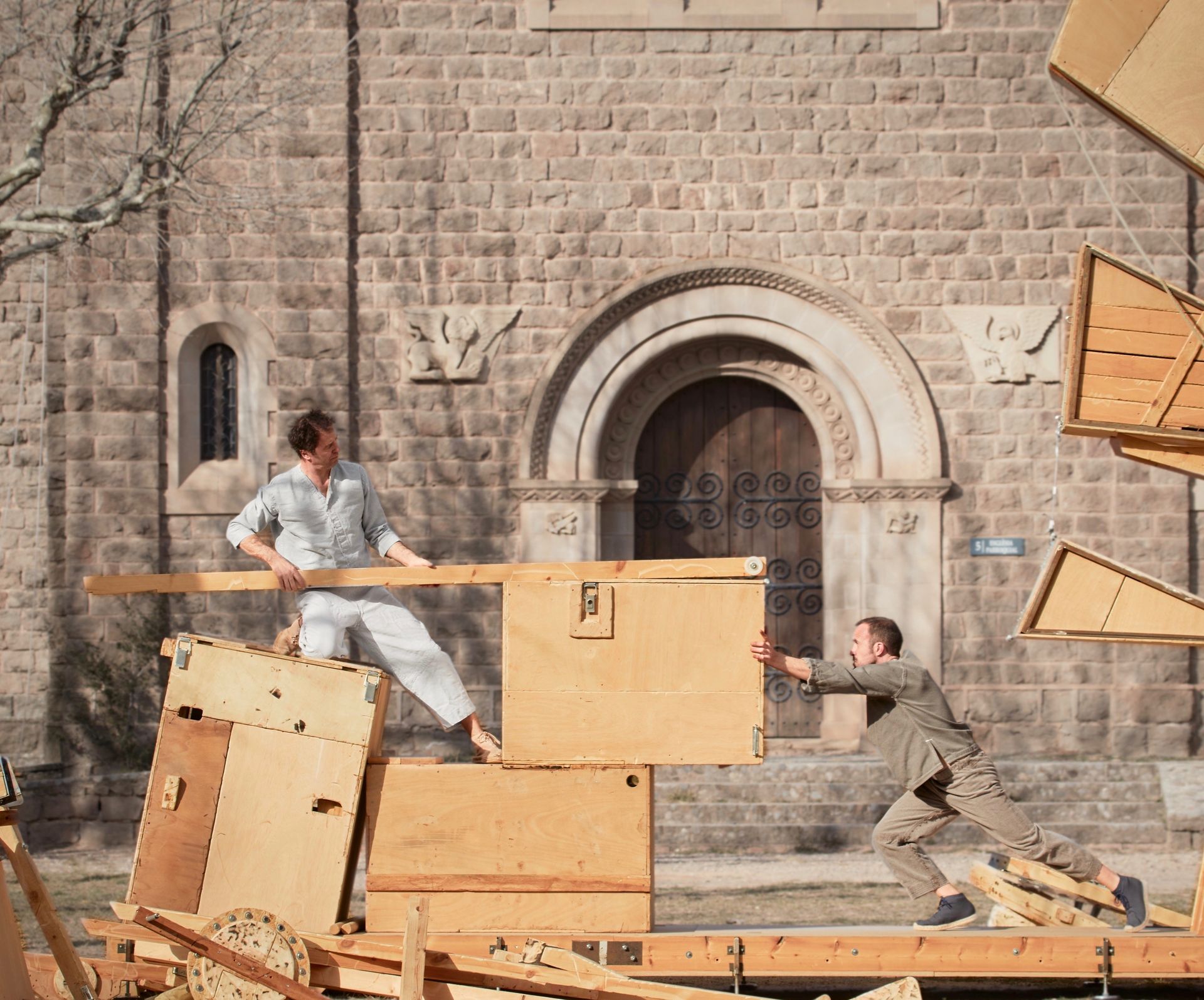 Farbfoto. Vor einem Gebäude aus hellem Gemäuer stapeln zwei Personen große Holzstücke und Holzboxen in eine Skulptur, auf der sie selbst stehen. Die Formen und Gegenstände ergeben keine klar bestimmbare Konstruktion.