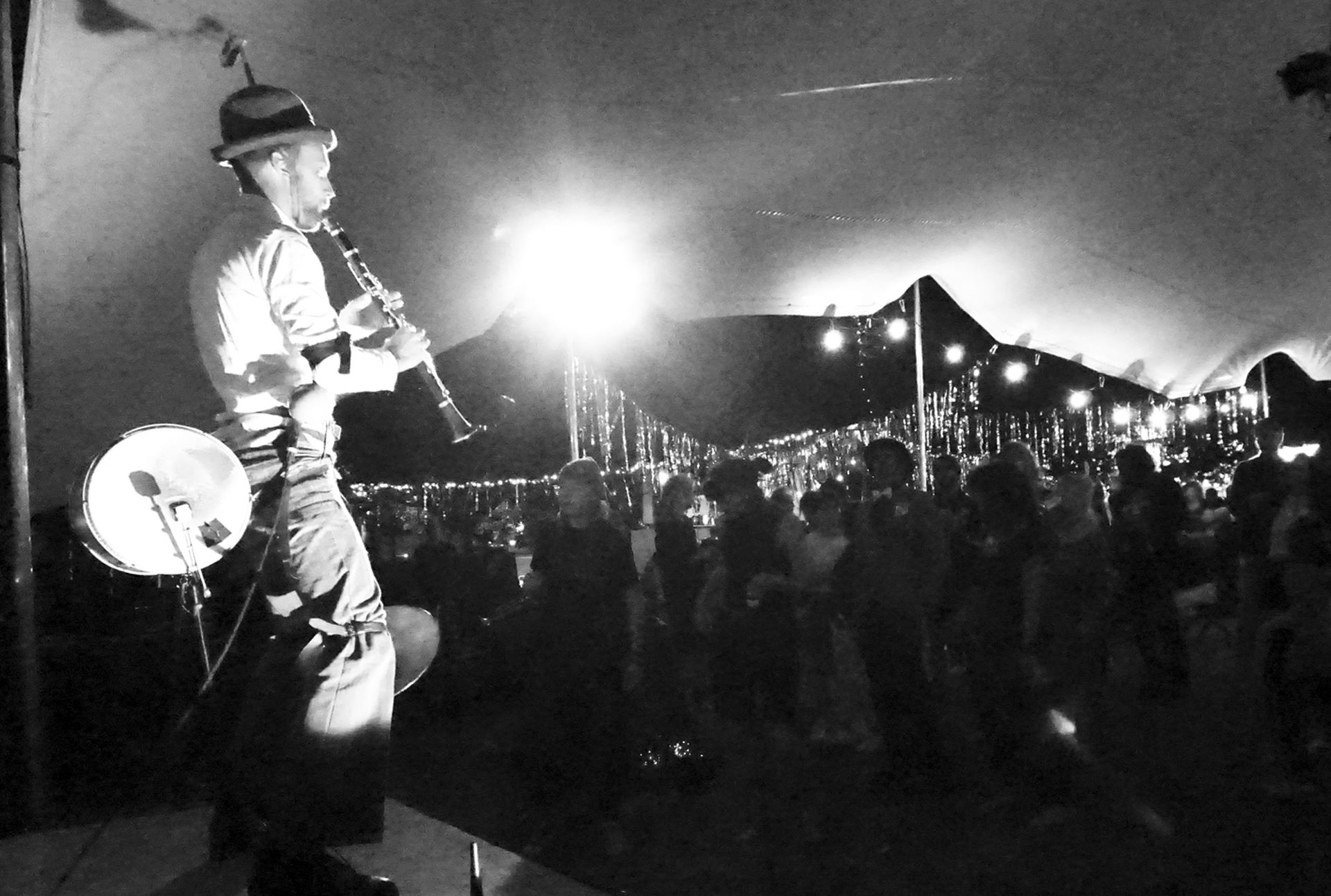Schwarz-weiß Foto. Martin Kaspar steht links im Bild, Klarinette spielend und mit einer Trommel. Rechts im Bild steht das Publikum. Das Konzert findet in einem Zelt statt, in dem sich einzelne Laternen befinden.