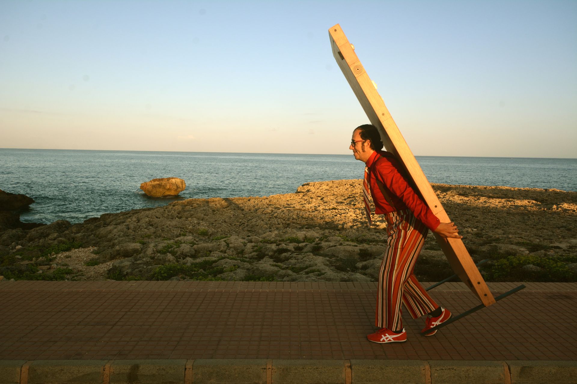 Farbfoto. Eine Person in rotem Hemd und rot-gestreifter Hose trägt eine hölzerne Tür vom rechten Rand kommend durch das Bild. Im Hintergrund sind ein Strand mit Gräsern und ein Meer zu sehen.