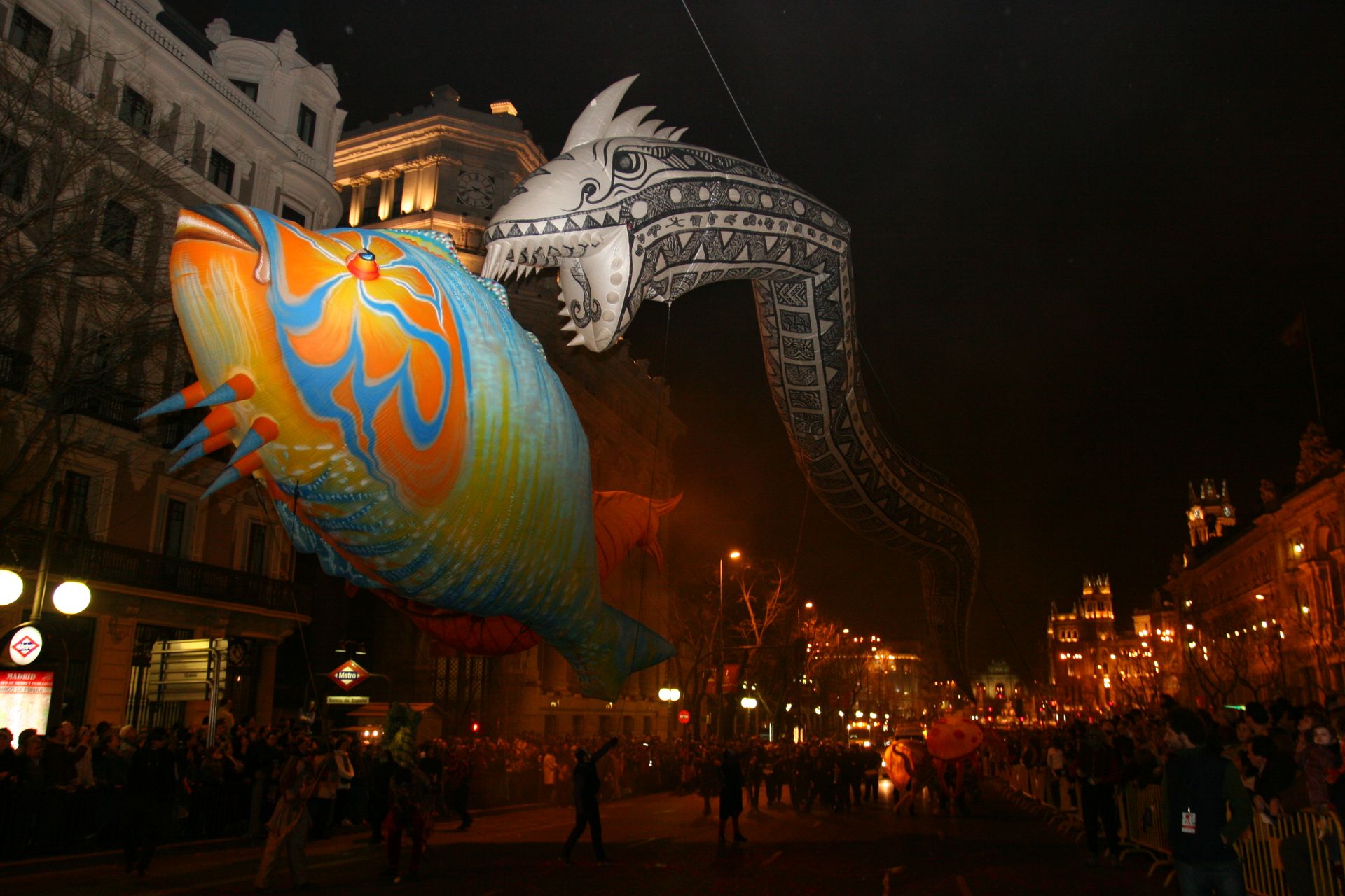Farbfoto. In einer dämmrig beleuchteten Straße, auf der Menschen stehen, fliegen zwei übergroße Ballons über ihre Köpfe hinweg. Ein Ballon sieht aus wie ein blau-oranger Fisch, der zweite ist ein schwarz-weißer Aal.