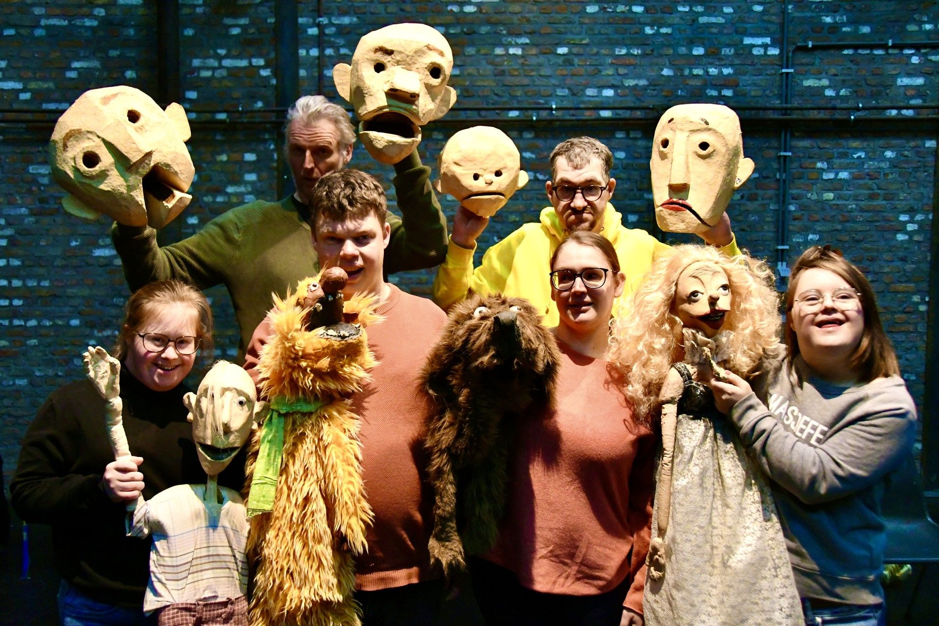 Farbfoto von sechs Personen vor einer blau angeleuchteten Wand. Alle halten Puppen aus Schaumstoff. Puppen von menschlichen Köpfen, die grimmig oder erschrocken schauen; Tier-Puppen mit braunem und orangem Fell; Mensch-Puppen mit Körperteilen.